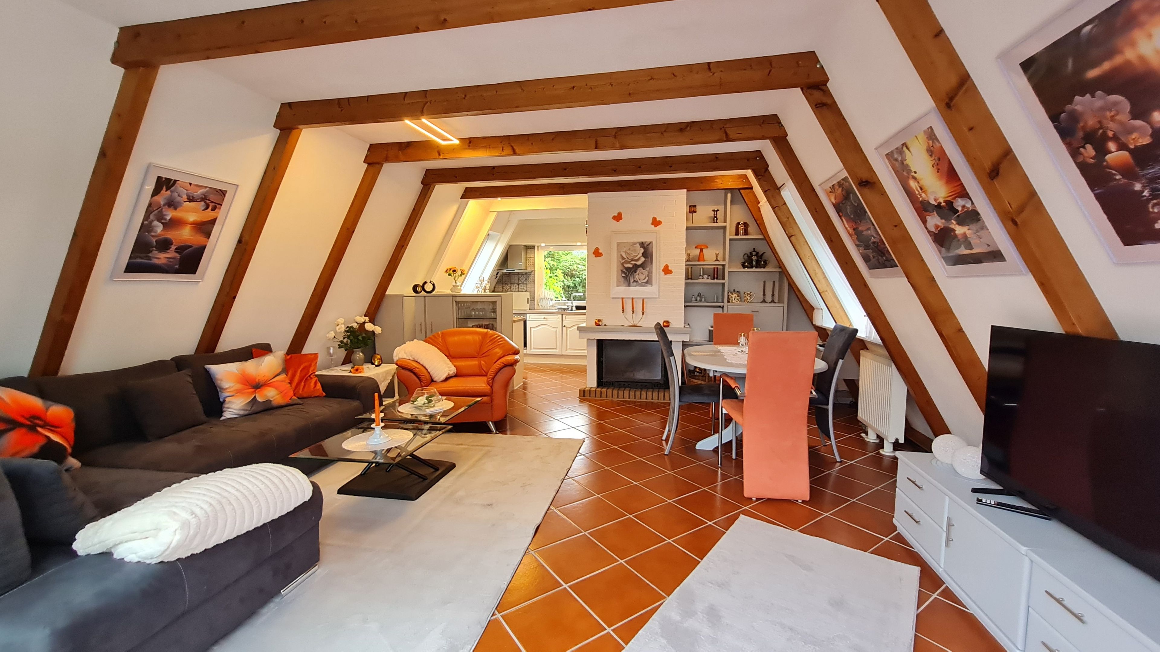 Ein gemütliches Wohnzimmer mit einer Couch, einem Couchtisch und orangefarbenen Sesseln. Im Hintergrund ist ein Essbereich mit Tisch und Stühlen zu sehen.