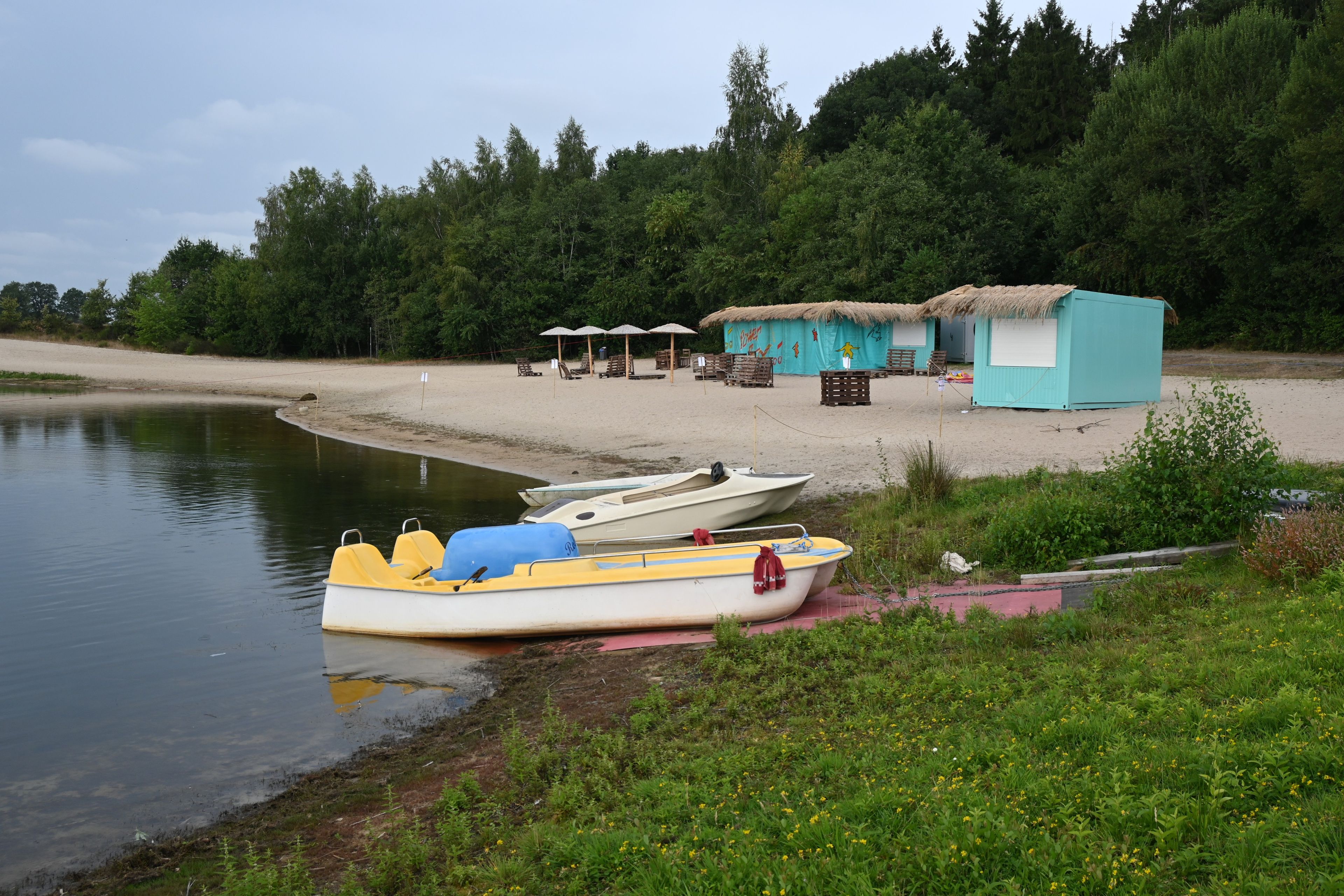 Eine Seenlandschaft mit einem am Ufer angelegtem Ruderboot, einer kleinen Hütte sowie einer Reihe von Liegestühlen und Sonnenschirmen.