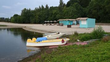 Eine Seenlandschaft mit einem am Ufer angelegtem Ruderboot, einer kleinen Hütte sowie einer Reihe von Liegestühlen und Sonnenschirmen.