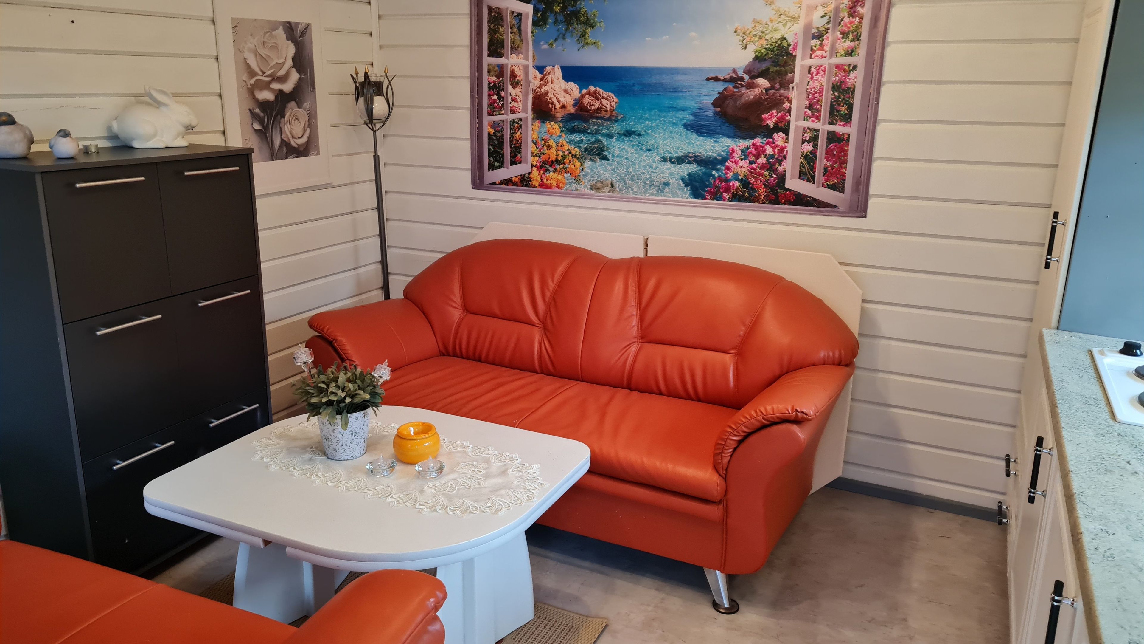 Ein Zimmer mit einem roten Sofa, einem weißen Tisch mit Vase und Kerze, einer schwarzen Kommode und einer Wand mit einem Landschaftsbild.