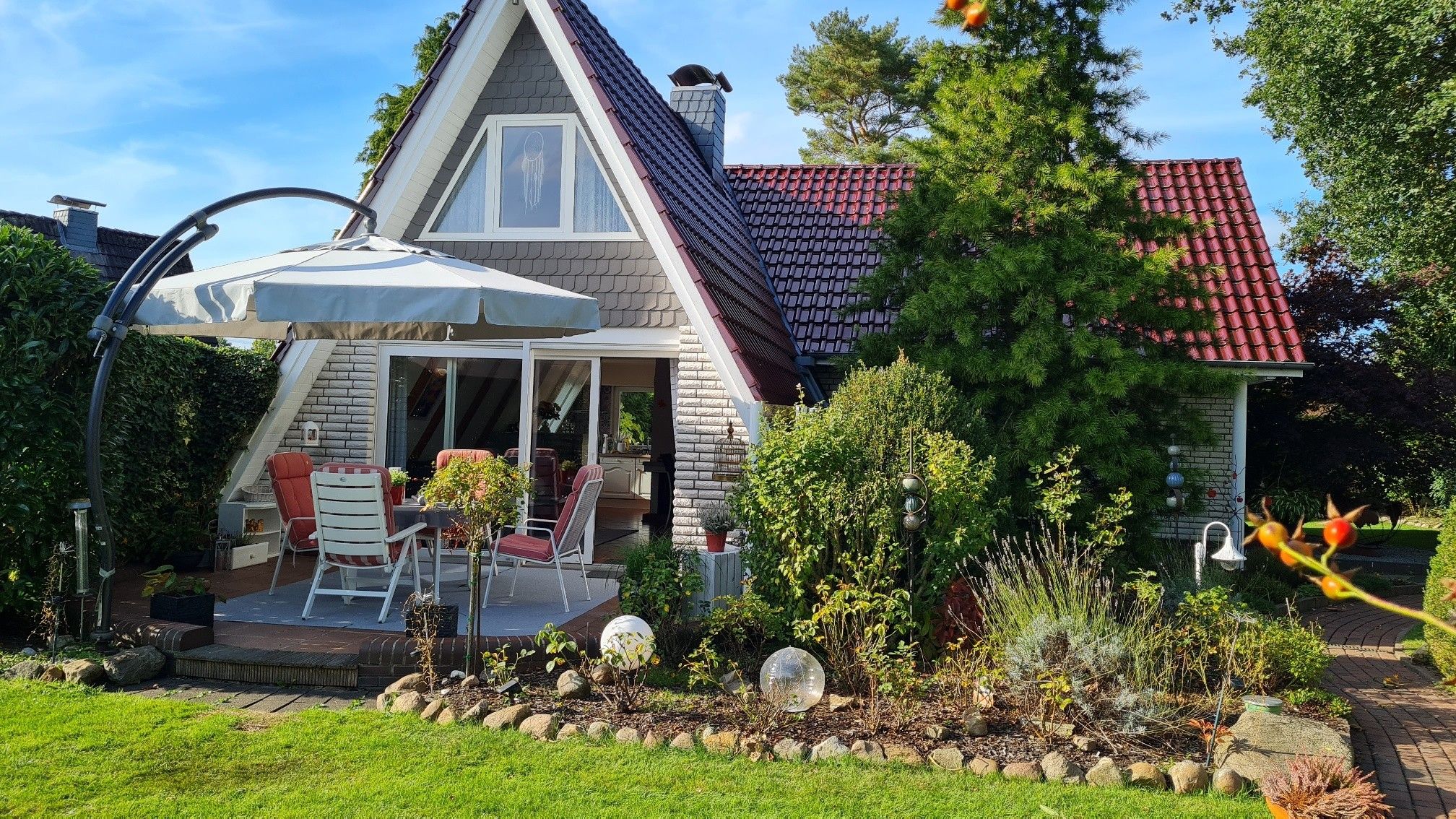 A house with a sloped roof, a covered patio with outdoor furniture, and a garden with various plants and decorative elements.