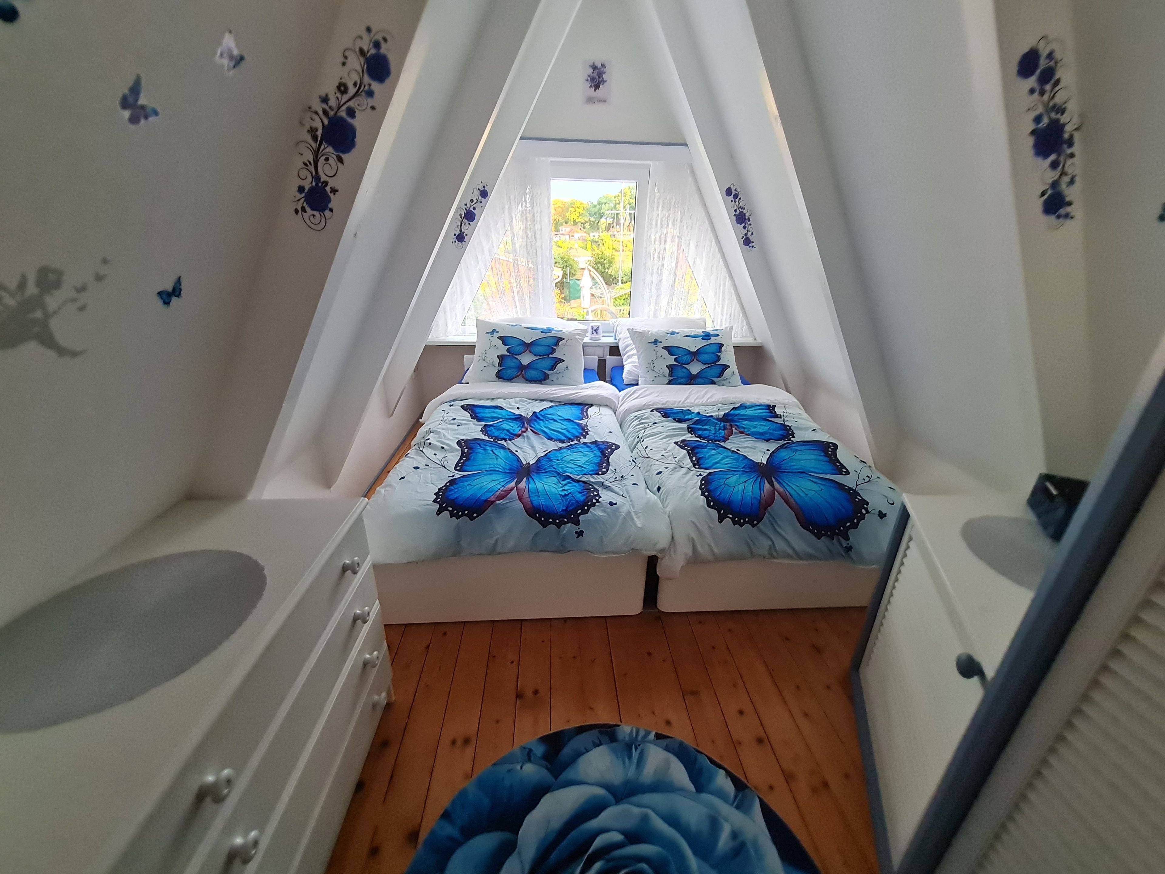 A bedroom with a sloped ceiling, a bed with butterfly-themed bedding, a window with a view of greenery, and a white dresser.