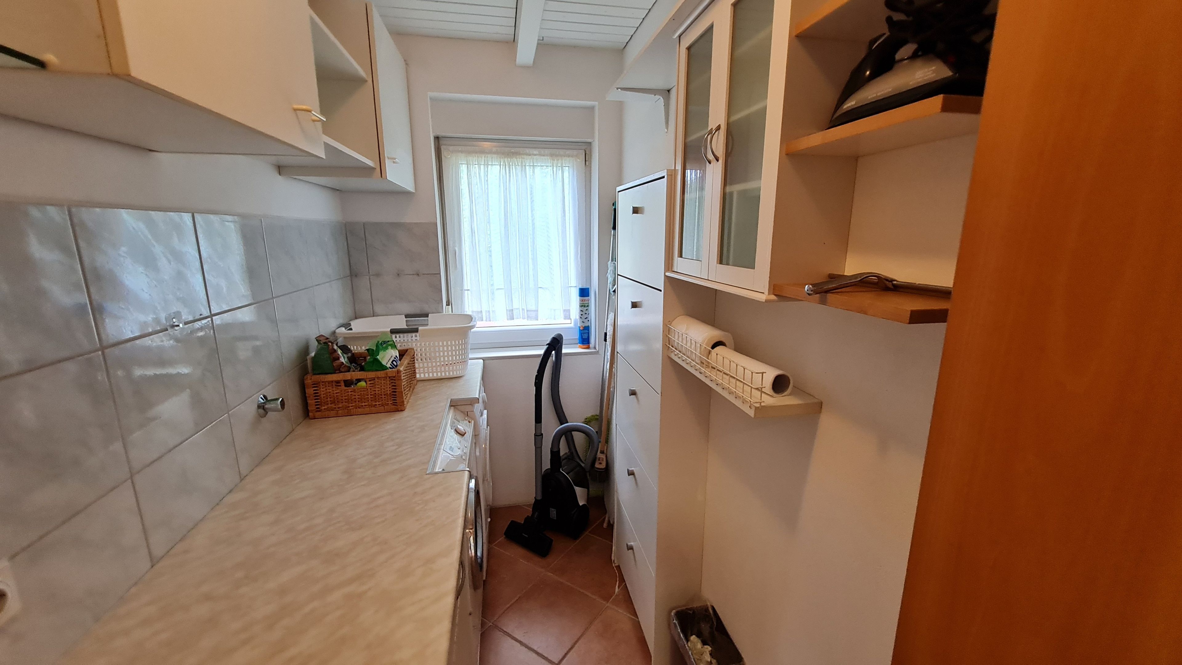 Eine schmale Küche mit gefliesten Wänden, einem Fenster mit Vorhängen, einem Kühlschrank und einem Staubsauger.