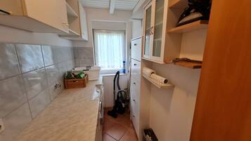 Eine schmale Küche mit gefliesten Wänden, einem Fenster mit Vorhängen, einem Kühlschrank und einem Staubsauger.