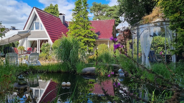 Uma casa com telhado vermelho, um lago com uma estátua e móveis de jardim.