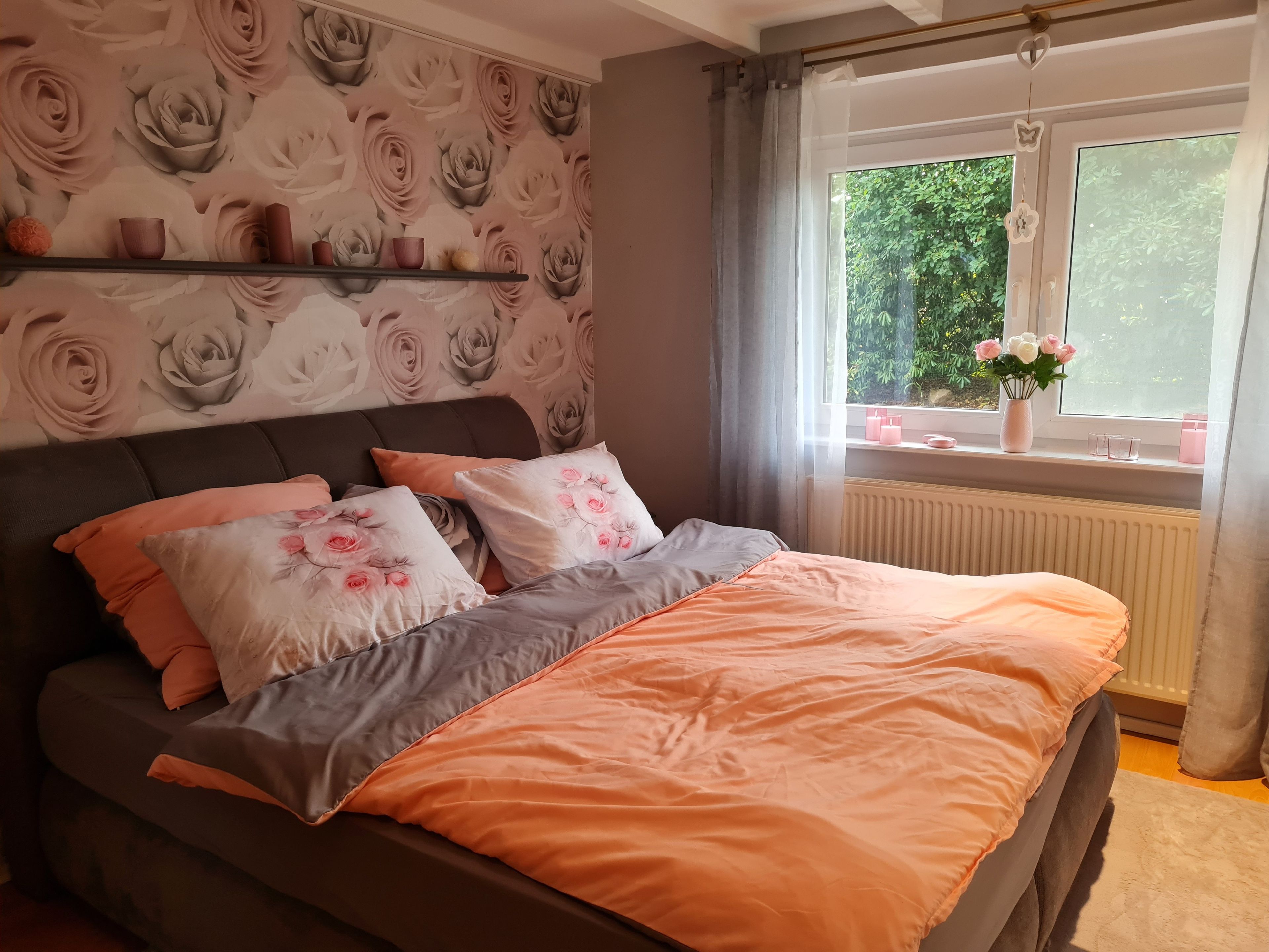Ein Schlafzimmer mit einem Bett, Kissen, einem Regal mit Dekoration, einem Fenster mit Blick auf Grünflächen und einer Heizung.