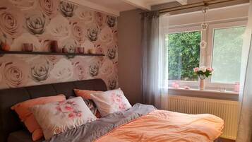 Ein Schlafzimmer mit einem Bett, Kissen, einem Regal mit Dekoration, einem Fenster mit Blick auf Grünflächen und einer Heizung.