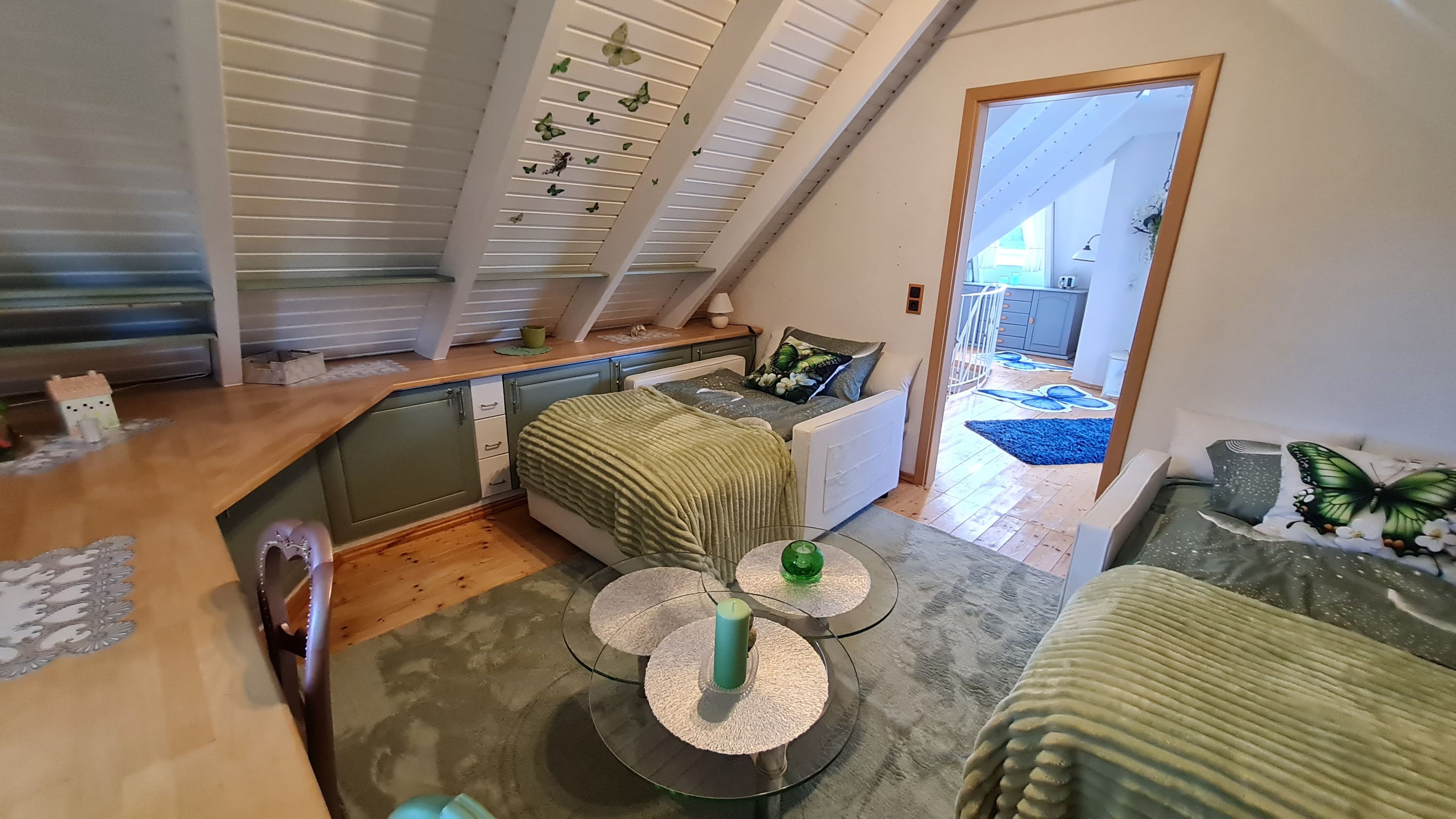 Ein gemütliches Schlafzimmer im Loft-Stil mit schrägem Dach, einem Bett mit grüner Decke, einem kleinen Tisch mit einer grünen Kerze und Blick in den Küchenbereich.
