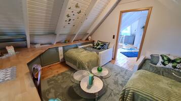 Ein gemütliches Schlafzimmer im Loft-Stil mit schrägem Dach, einem Bett mit grüner Decke, einem kleinen Tisch mit einer grünen Kerze und Blick in den Küchenbereich.