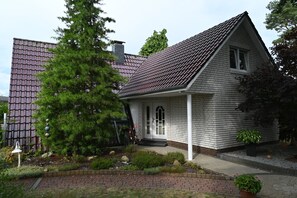 Uma casa térrea com telhado inclinado, varanda frontal e um jardim bem cuidado, com diversas plantas e uma grande árvore perene.