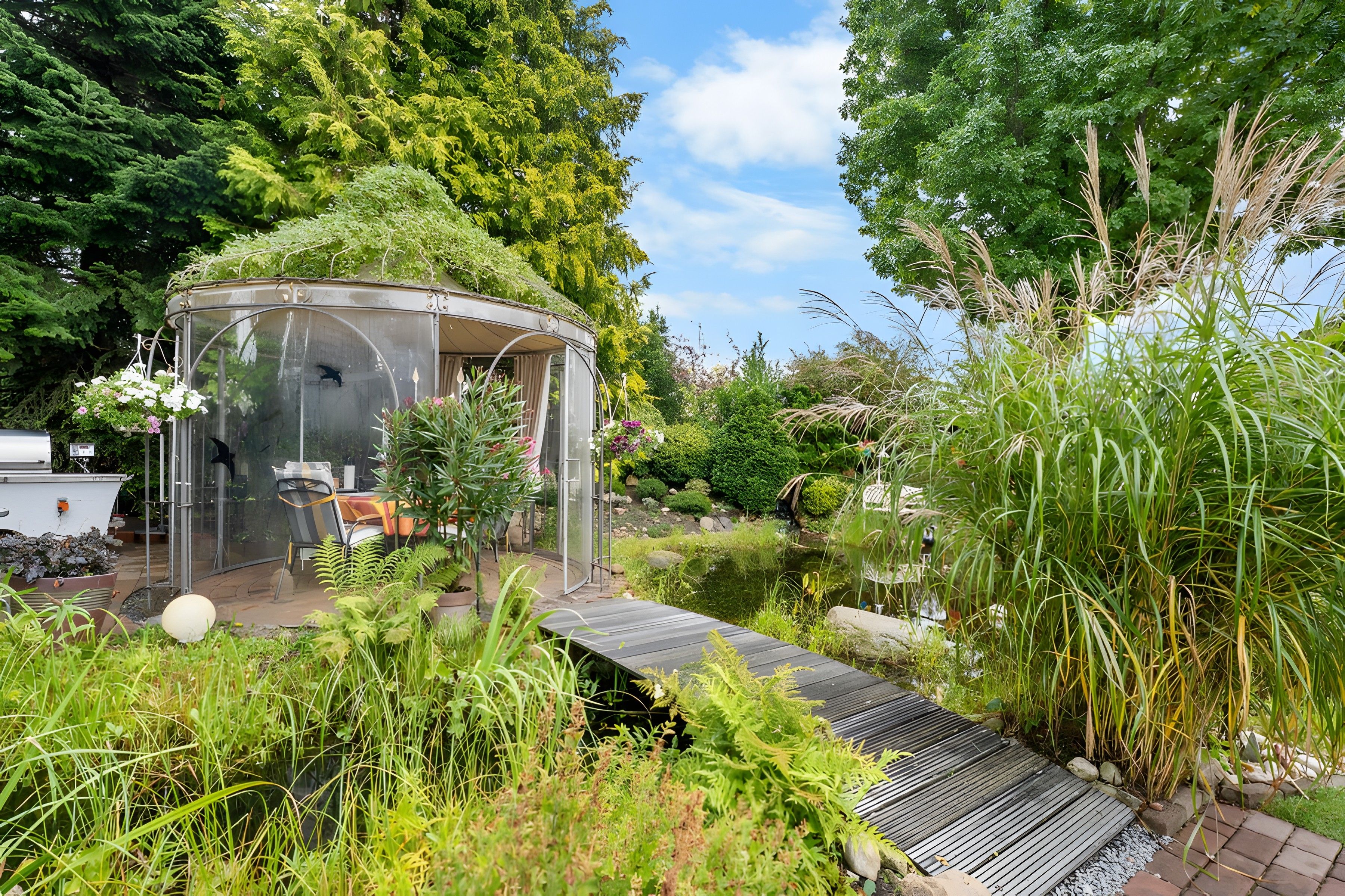 A glass-enclosed gazebo with a table and chairs, surrounded by lush greenery and a small pond.