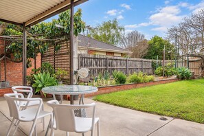 Un espace patio avec une table ronde en verre et des chaises blanches, entouré d’une clôture en bois et de diverses plantes.