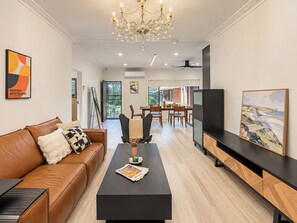 A modern living room with a brown leather sofa, a black coffee table with magazines, a chandelier, and artwork on the walls. - Cozy Bentleigh Family Retreat (Bentleigh)
