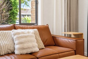 Ein braunes Ledersofa mit zwei Kissen, ein kleiner Holztisch und ein Fenster mit Blick auf Grünflächen.