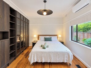 A bedroom with a large bed, wooden flooring, a wardrobe, a window with a view of greenery, and a ceiling light. - Cozy Bentleigh Family Retreat (Bentleigh)