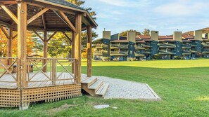 A wooden gazebo with a ramp, a grassy lawn, and a row of multi-story buildings with balconies. - Smugglers Notch | Snowy Retreat | Ski, Pools & Mountain Fun (Jeffersonville)