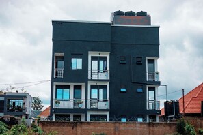 A multi-story building with balconies, large windows, and a brick wall in the foreground. - Mutoni Teddy (Kigali)