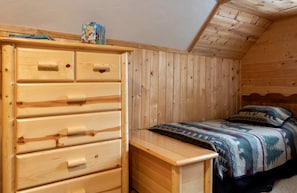 A bedroom with a wooden dresser, a bed with patterned bedding, and a sloped ceiling. - Adventurous Cabin with Hot Tub & Fire Pit for Relaxing in Island Park, Idaho (Island Park)