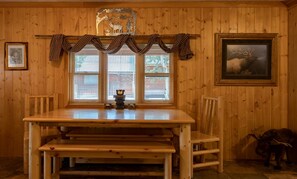A wooden interior with a dining table, chairs, a window with blinds, a framed picture, and a bear figurine. - Adventurous Cabin with Hot Tub & Fire Pit for Relaxing in Island Park, Idaho (Island Park)