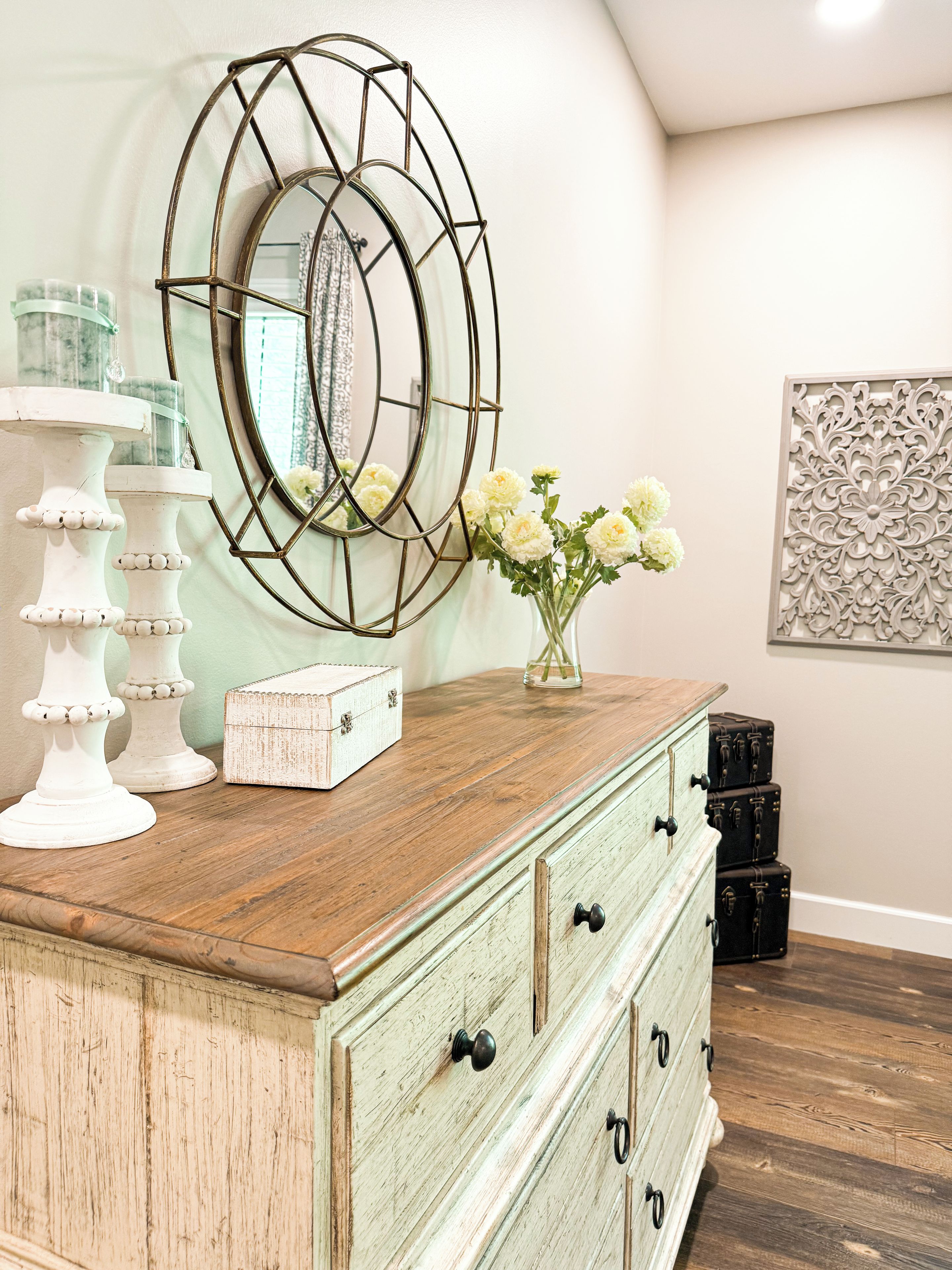 Een houten dressoir met laden, een spiegel met geometrisch lijstwerk, een vaas met bloemen en een decoratief object aan de muur.