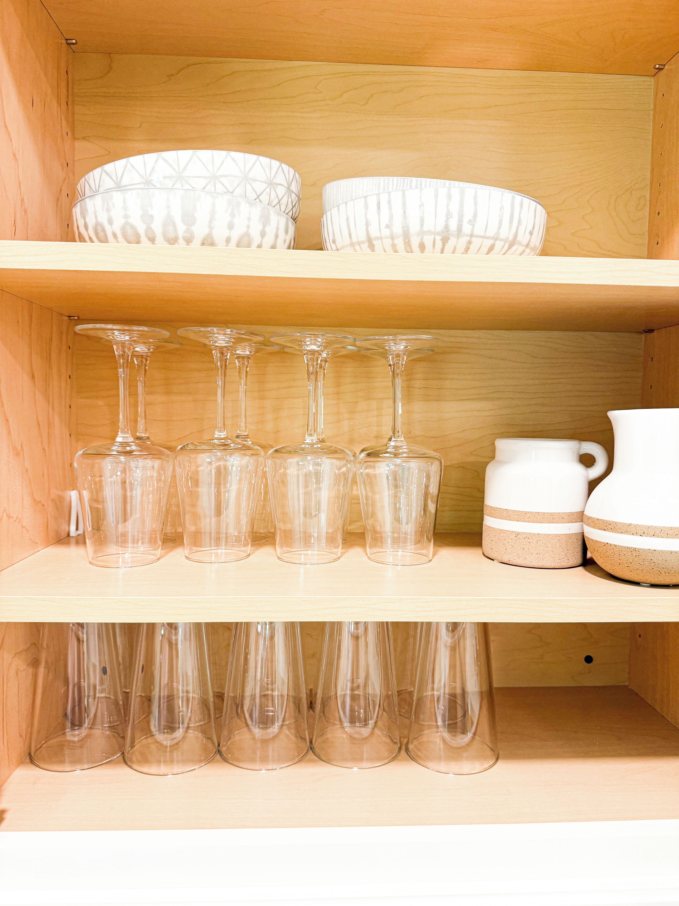 Une étagère en bois sur laquelle sont posés des verres à vin en verre transparent, un pichet en céramique blanche et deux bols à motifs.