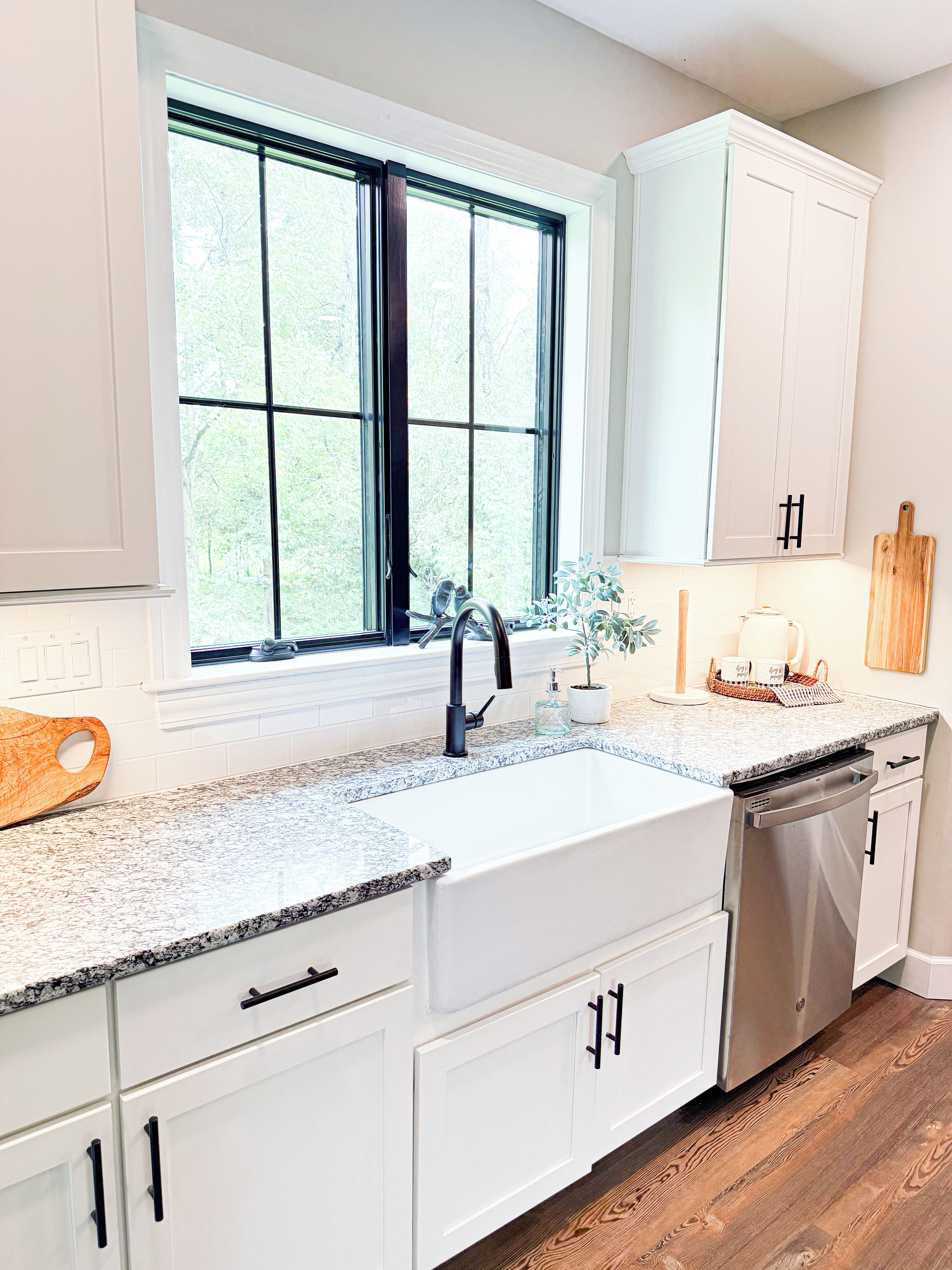 Una cocina con muebles blancos, un lavamanos doble y una ventana con vistas a árboles.