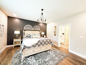 A bedroom with a large bed, two bedside tables, a chandelier, and a door leading to another room.