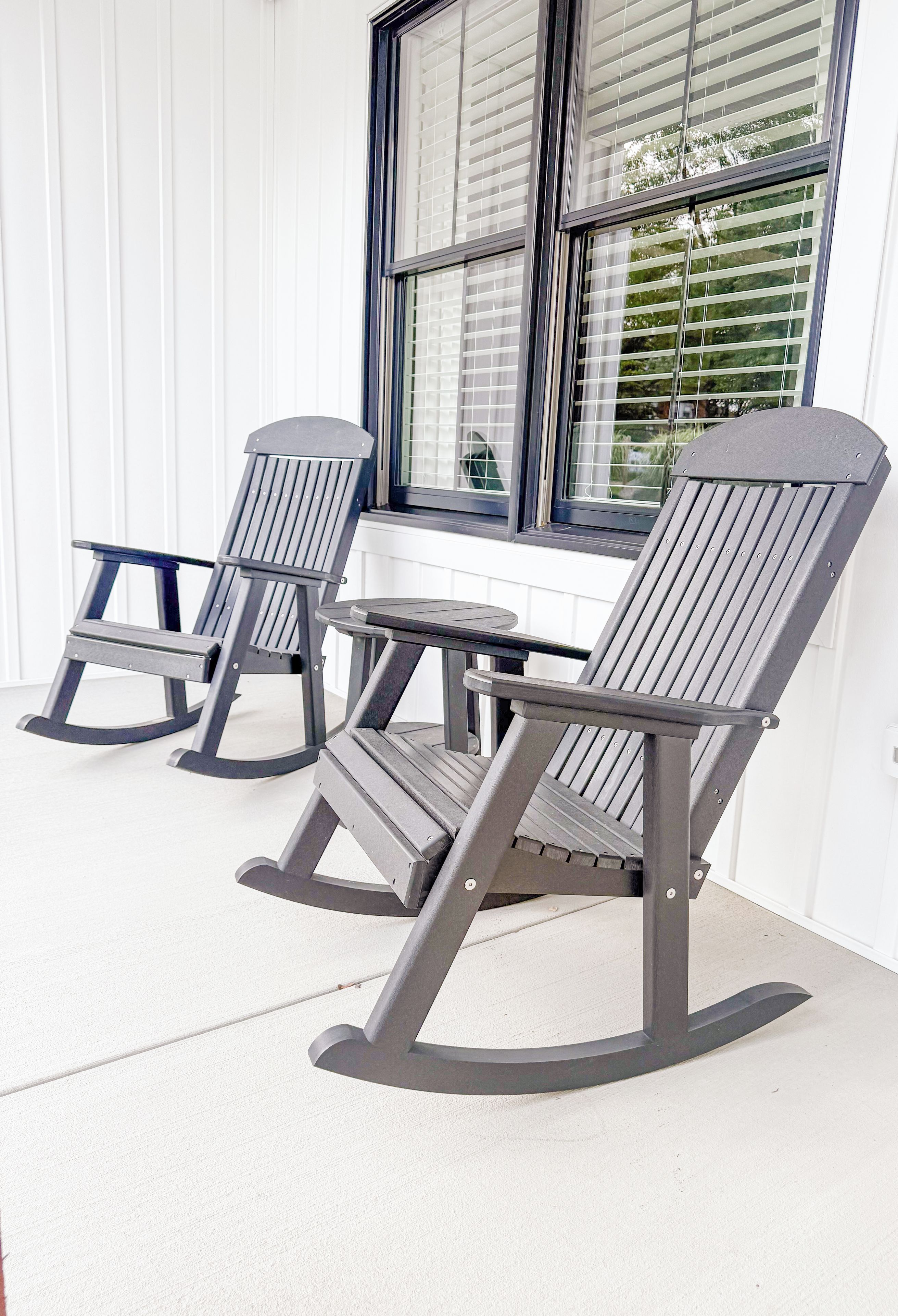 Three rocking chairs with a table on a porch with a window and blinds.