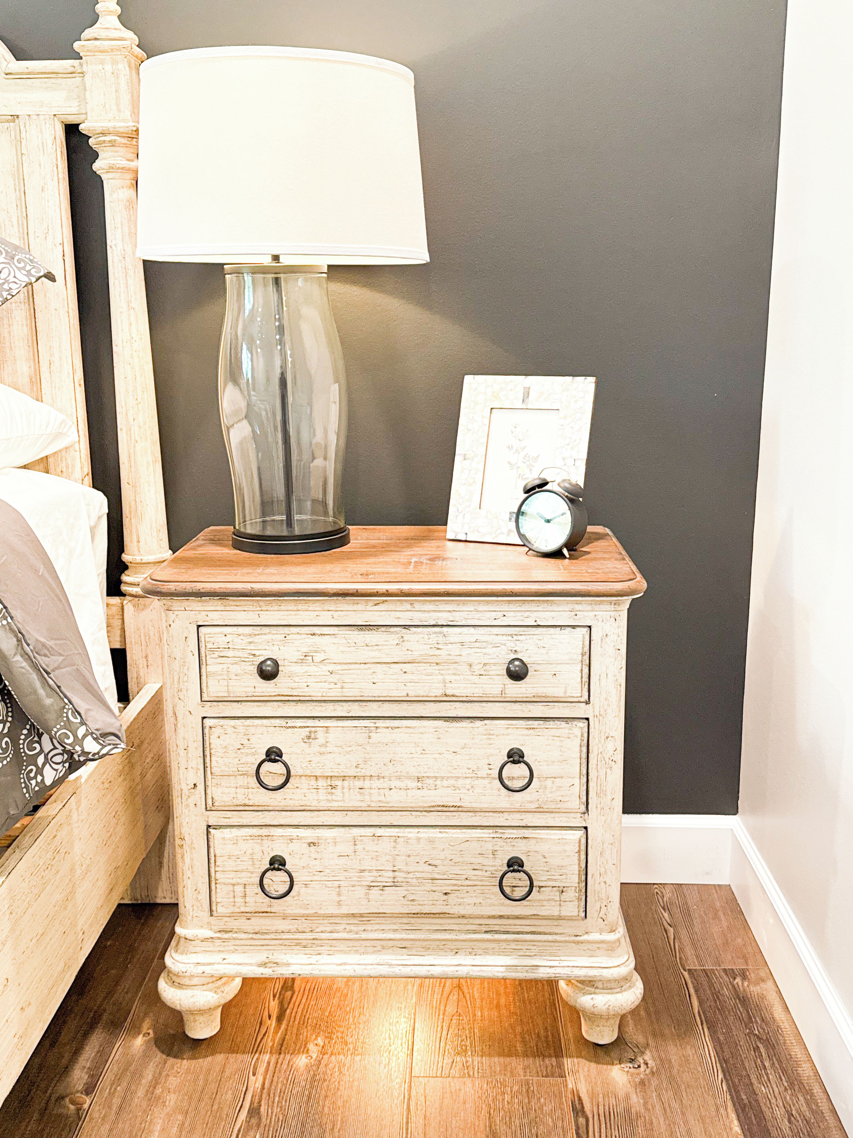 A wooden nightstand with a lamp, a framed picture, and an alarm clock.