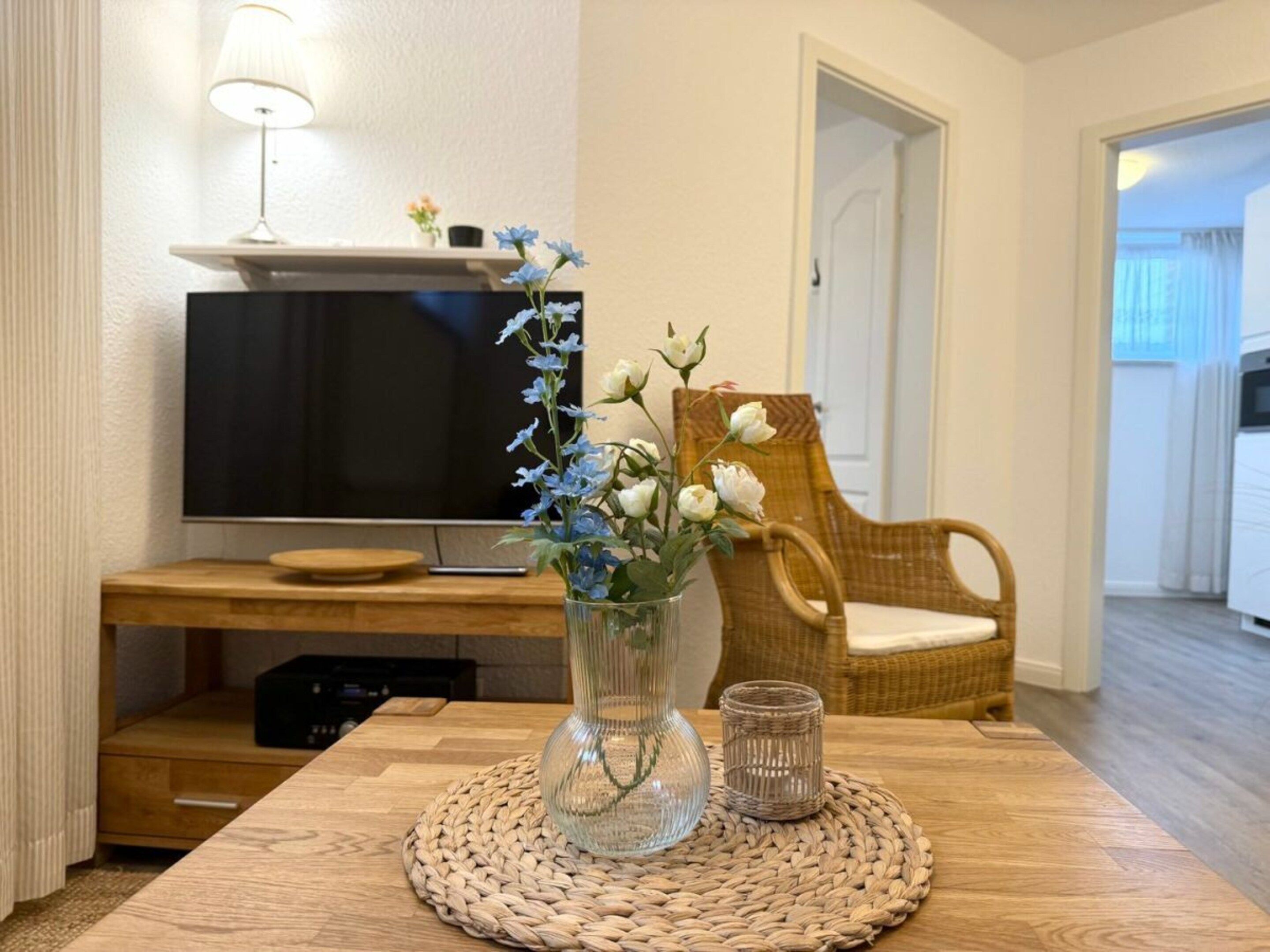 Et værelse med et træbord, en glasvase med blomster, en fletstol, et fjernsyn og en vævet underlag.