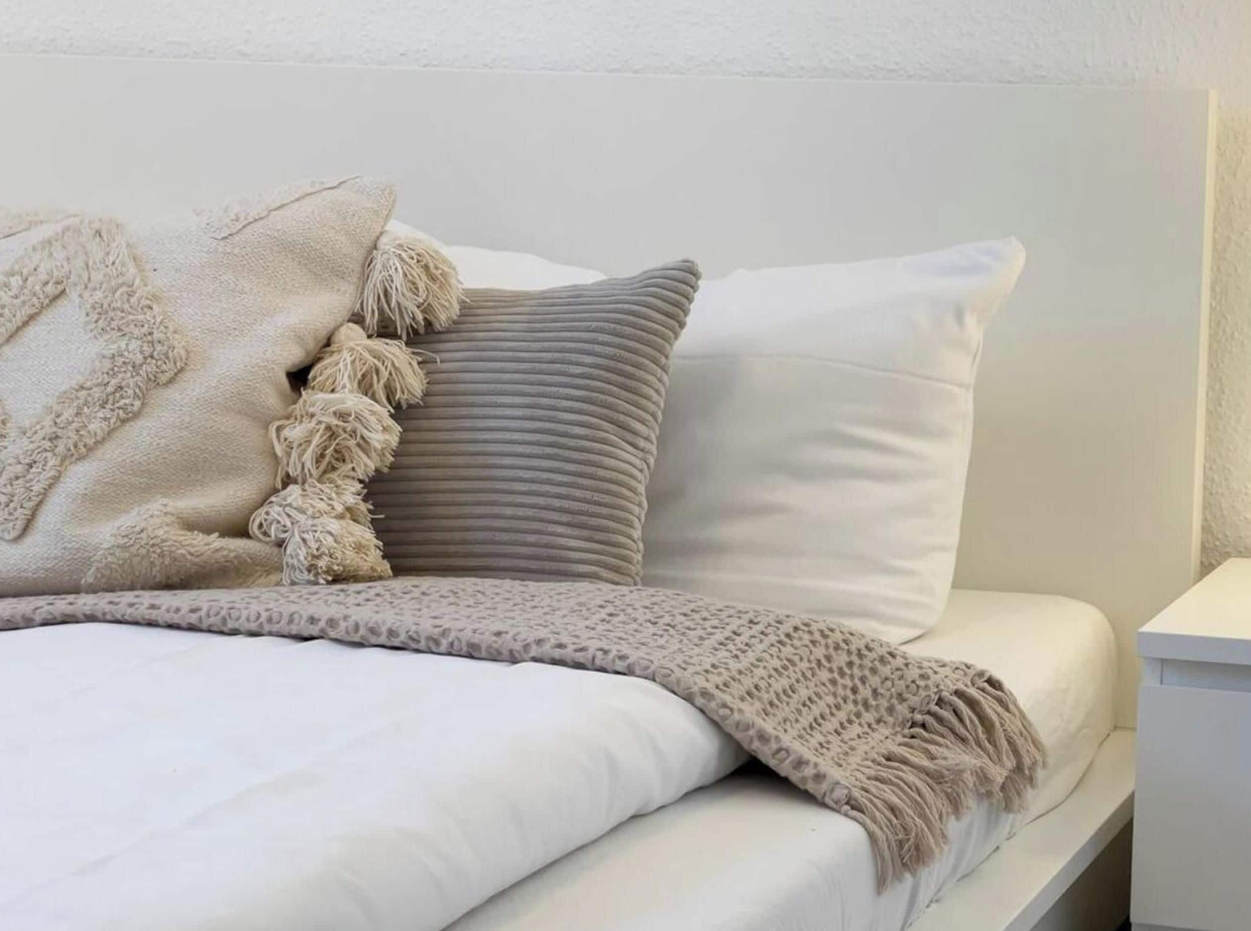 A bed with a white mattress, a grey pillow, and a beige throw blanket.