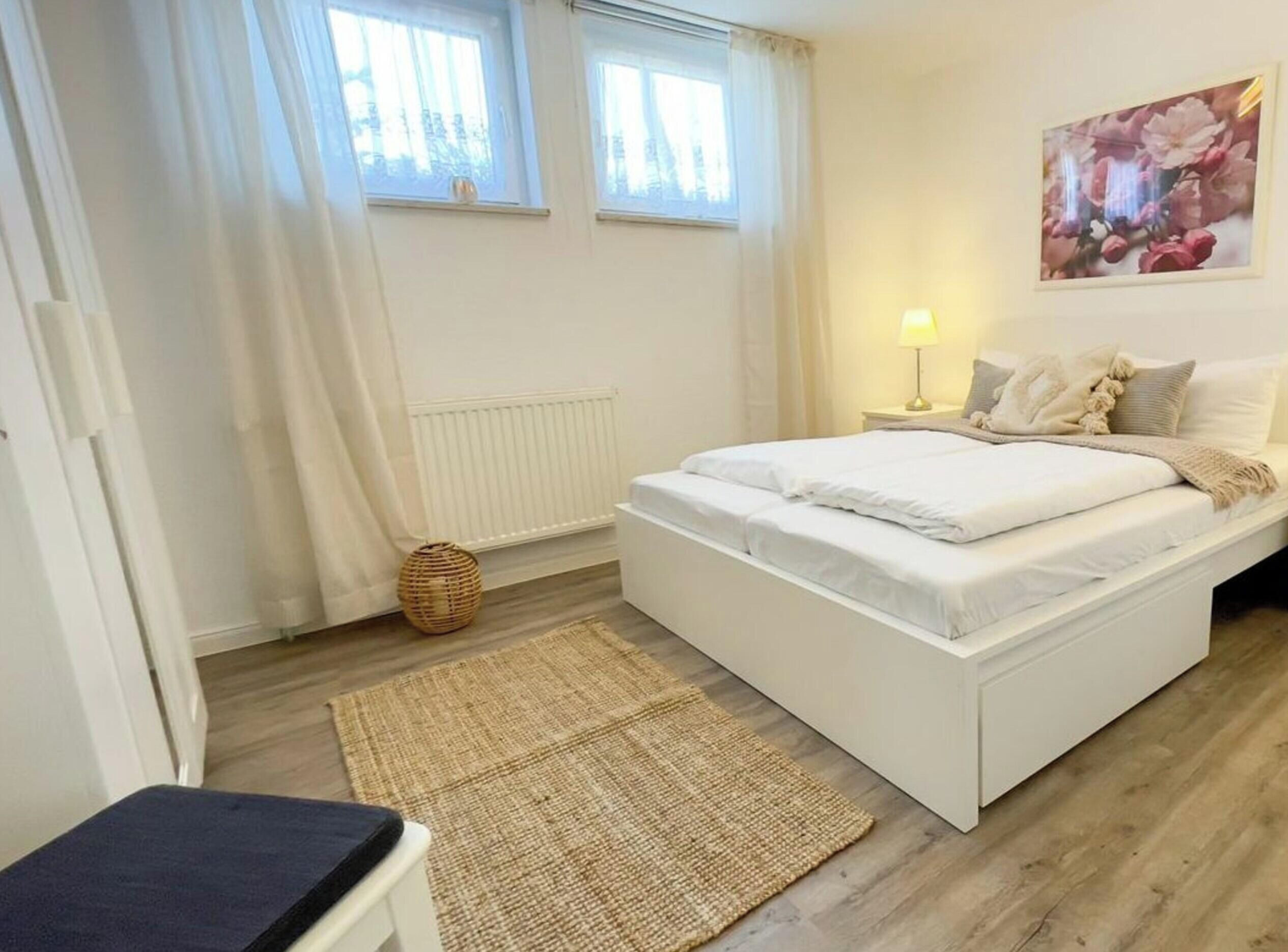 Et soveværelse med en seng, et natbord med lampe, et vindue og et billede hængt på væggen.