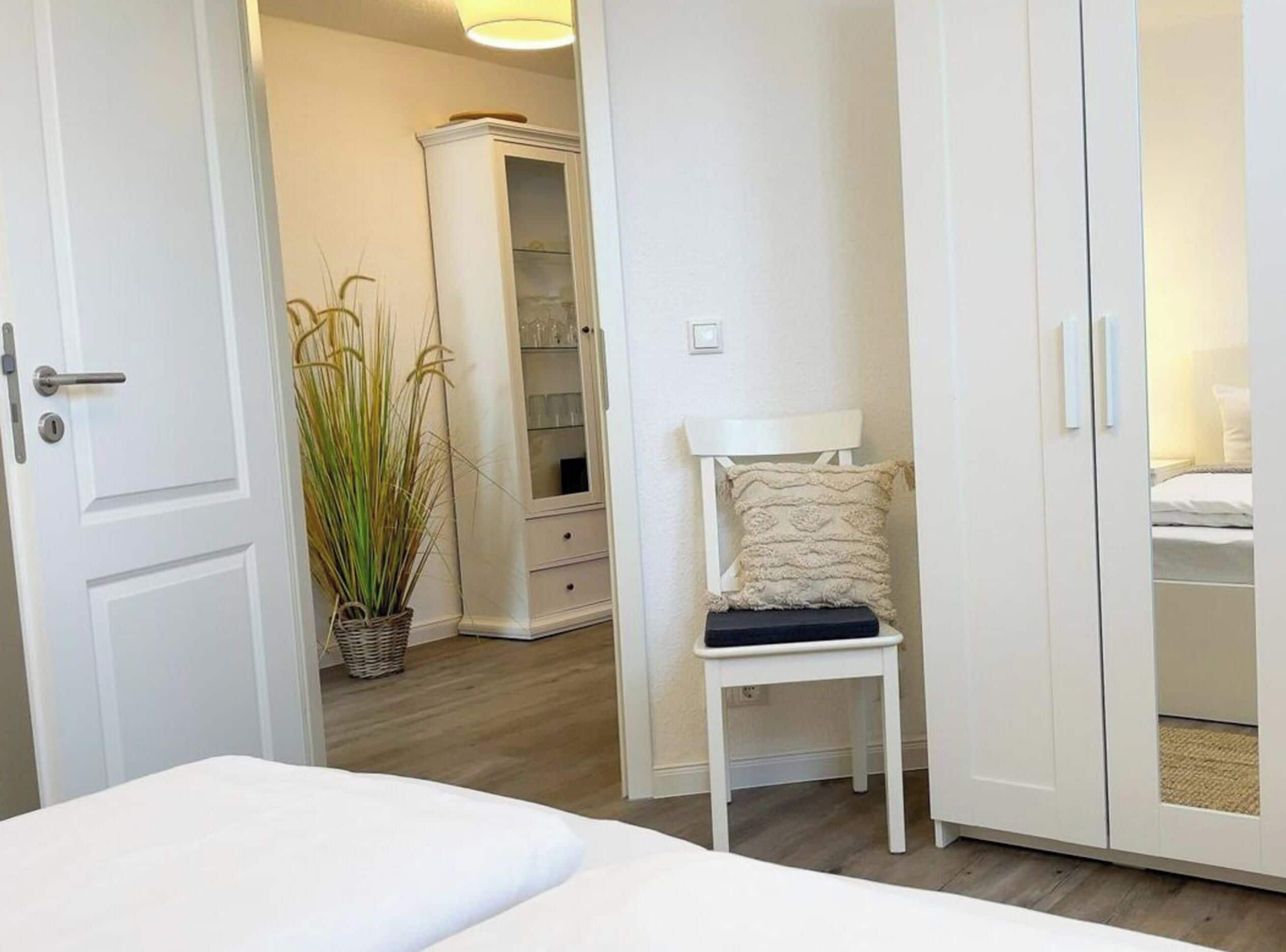Un dormitorio con una cama, un armario blanco, una mesita de noche con un almohadón y una planta.