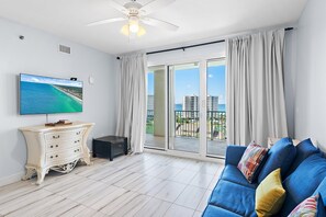 A modern living room with a flat-screen TV, a white dresser, a blue sofa with colorful cushions, and a balcony with a view of buildings and the sea. - Ariel Dunes II 909 (Miramar Beach)