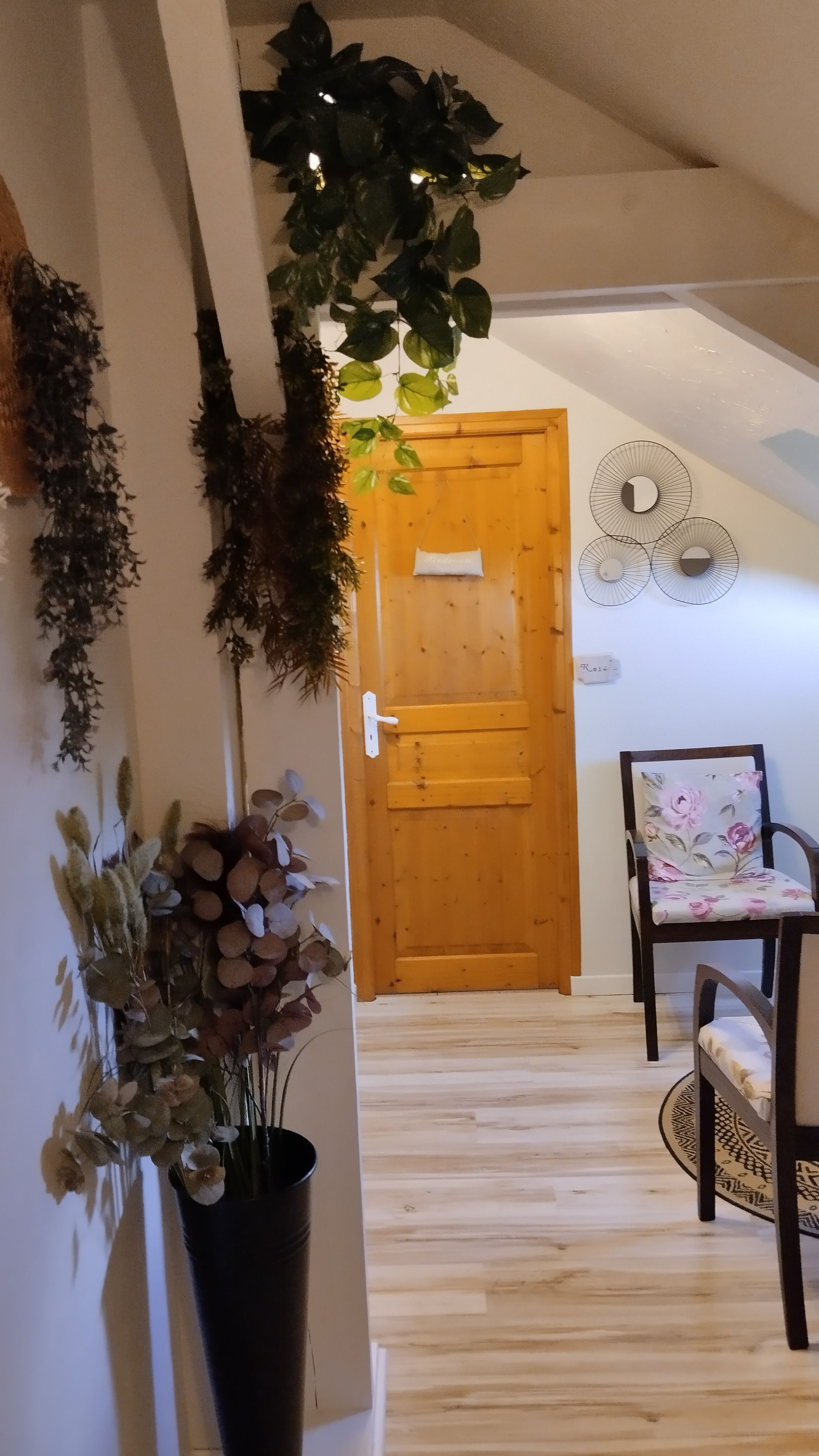 A hallway with a wooden door, a chair with floral upholstery, and a potted plant with dried flowers.