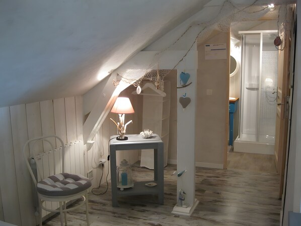 Un dormitorio acogedor en un ático, con techo inclinado, una mesita de noche con lámpara, una silla y una pequeña mesa con objetos decorativos.