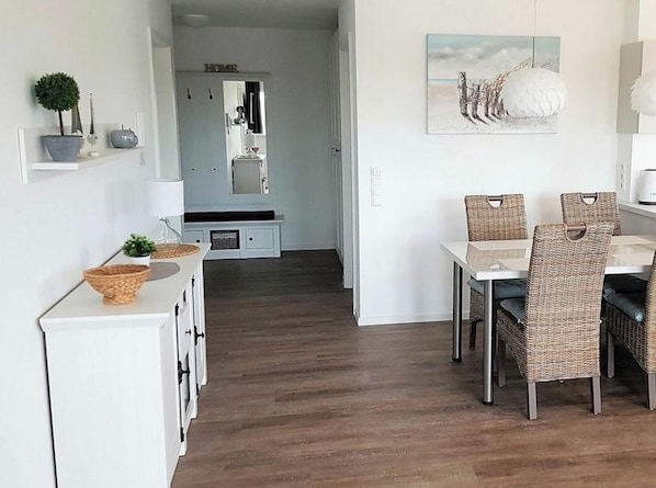 A dining area with wicker chairs and a white table, a kitchen with a white cabinet and a mirror, and a hallway leading to a bedroom. - 47.1.n Waterkant (Wangerland)