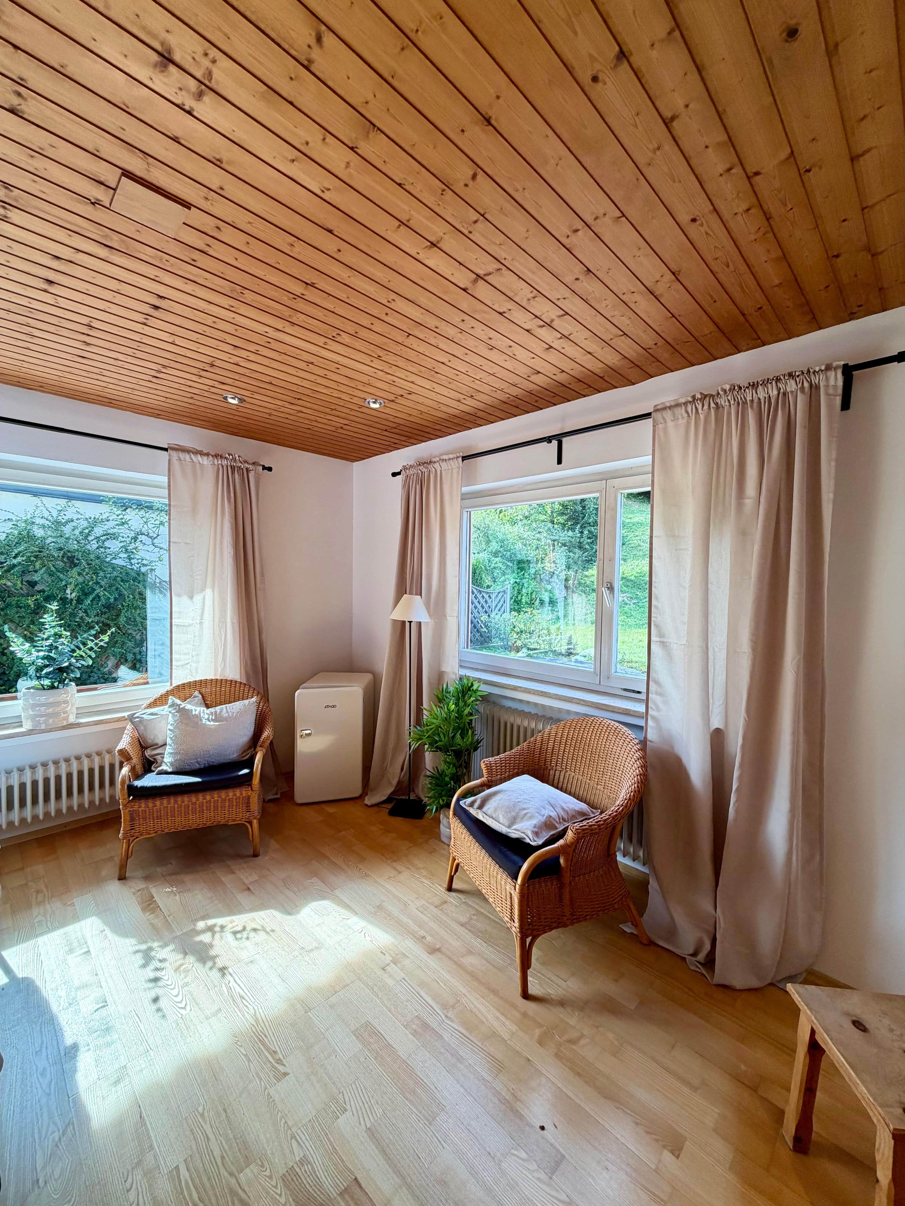 Ein Zimmer mit Holzboden, zwei Korbstühle mit Kissen, einem Heizkörper, einer Stehlampe und einem fenster mit Vorhängen.