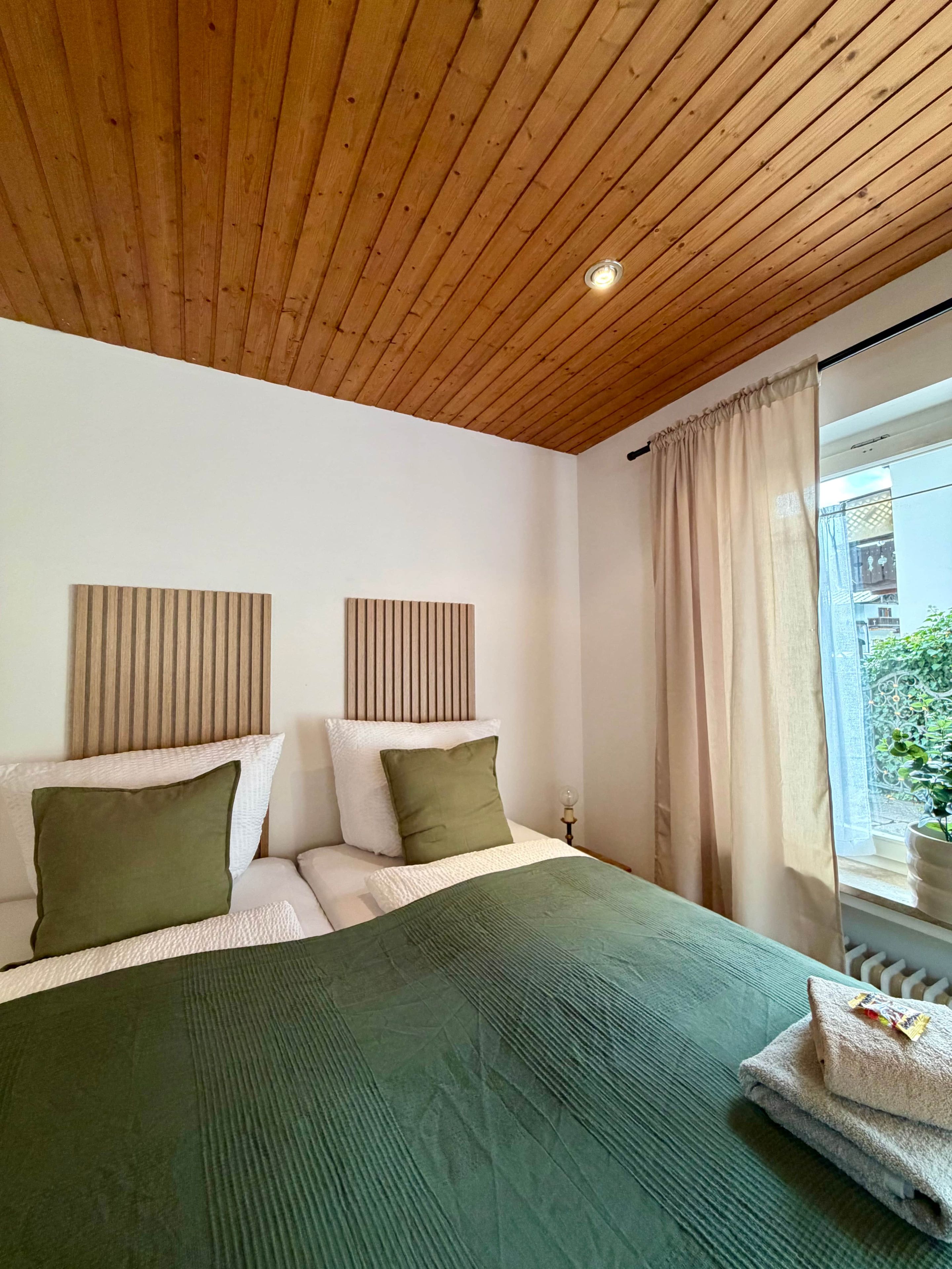 Een slaapkamer met een houten plafond, een bed met groen en wit beddengoed, een raam met gordijnen en uitzicht op groen.