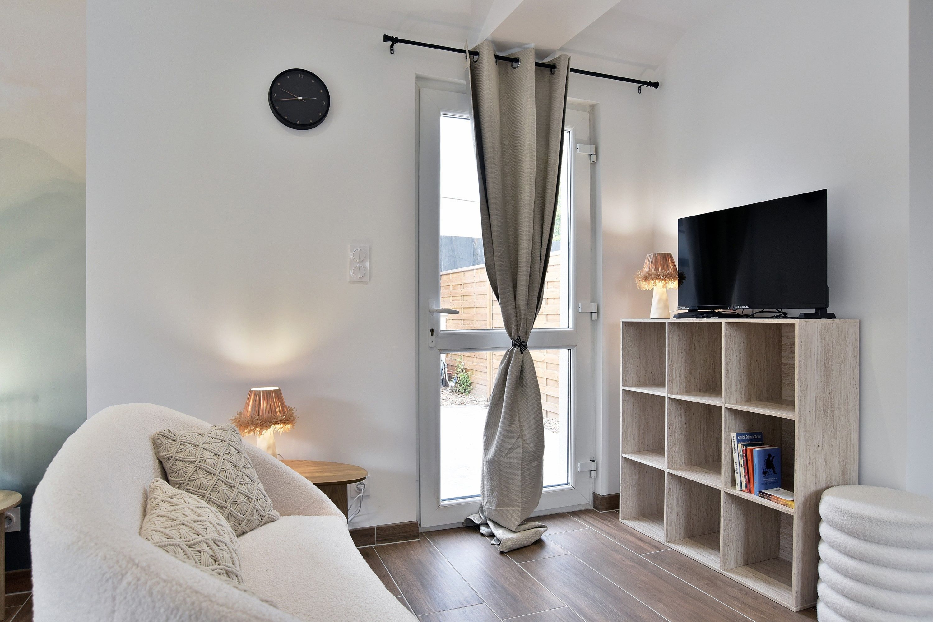 Un salon moderne avec un canapé, un téléviseur posé sur une étagère en bois et une fenêtre avec des rideaux.