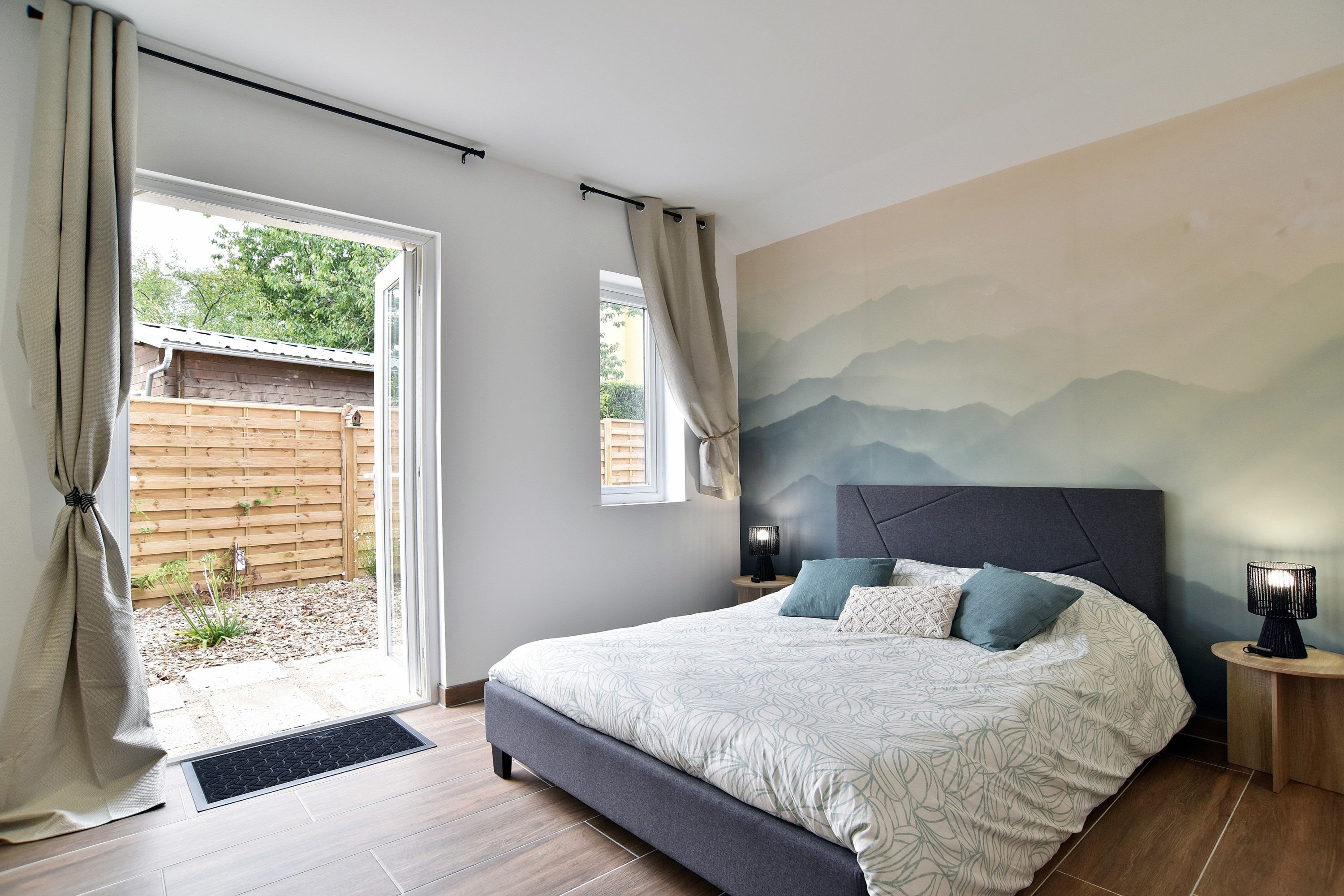 Une chambre à coucher comprenant un lit, une table de chevet, une lampe fixée au mur et une vue sur l’extérieur grâce à une porte coulissante.