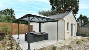 Ein überdachter Patio-Bereich mit Tisch und Stühlen, umgeben von einem Holenzaun und verschiedenen Pflanzen.
