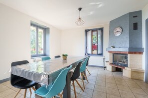A dining area with a table covered in plastic, blue chairs, a potted plant, and a fireplace with a clock on the wall. - Les Souvenirs du Reucy - vue montagne (La Bresse)