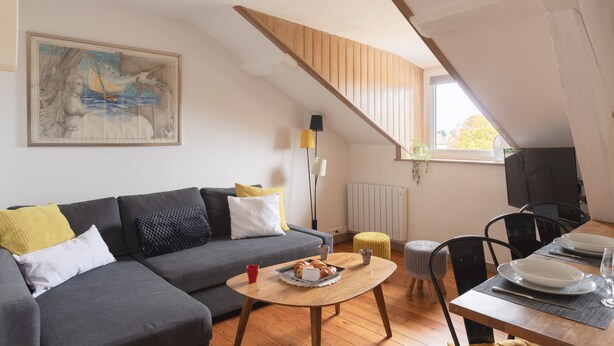 Un salon chaleureux avec un canapé gris, une table basse en bois et un coin repas avec une table dressée pour deux personnes.