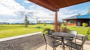 Eine Terrasse im Freien mit rundem Tisch und Stühlen, einem Picknicktisch und Blick auf ein grünes Feld und entfernte Gebäude.