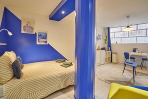 Ein Zimmer mit einem Bett, einem Schreibtisch und einem Stuhl. Es gibt ein Fenster mit Vorhängen, eine Lampe und ein Bild an der Wand.