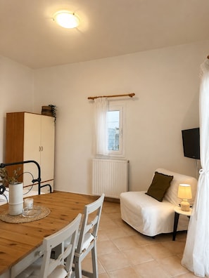 A room with a dining table, chairs, a cabinet, a radiator, a window with curtains, a sofa, a lamp, and a television.