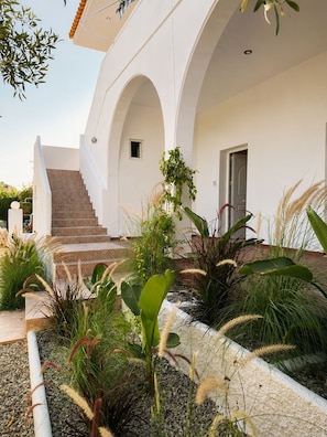 A white building with a staircase leading to an entrance, surrounded by various plants and a gravel pathway.
