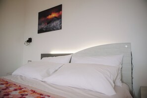 Ein Bett mit weißer Bettwäsche und einer floral gemusterten Decke, ein Wandbild mit einem Vulkans und ein schlichter Nachttisch mit einer Lampe.