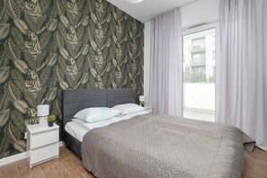 A bedroom with a bed, a nightstand, a lamp, and a window with curtains. - Łódź | Karola Borowieckiego 1 | Spacious Apartment | Terrace (Łódź)